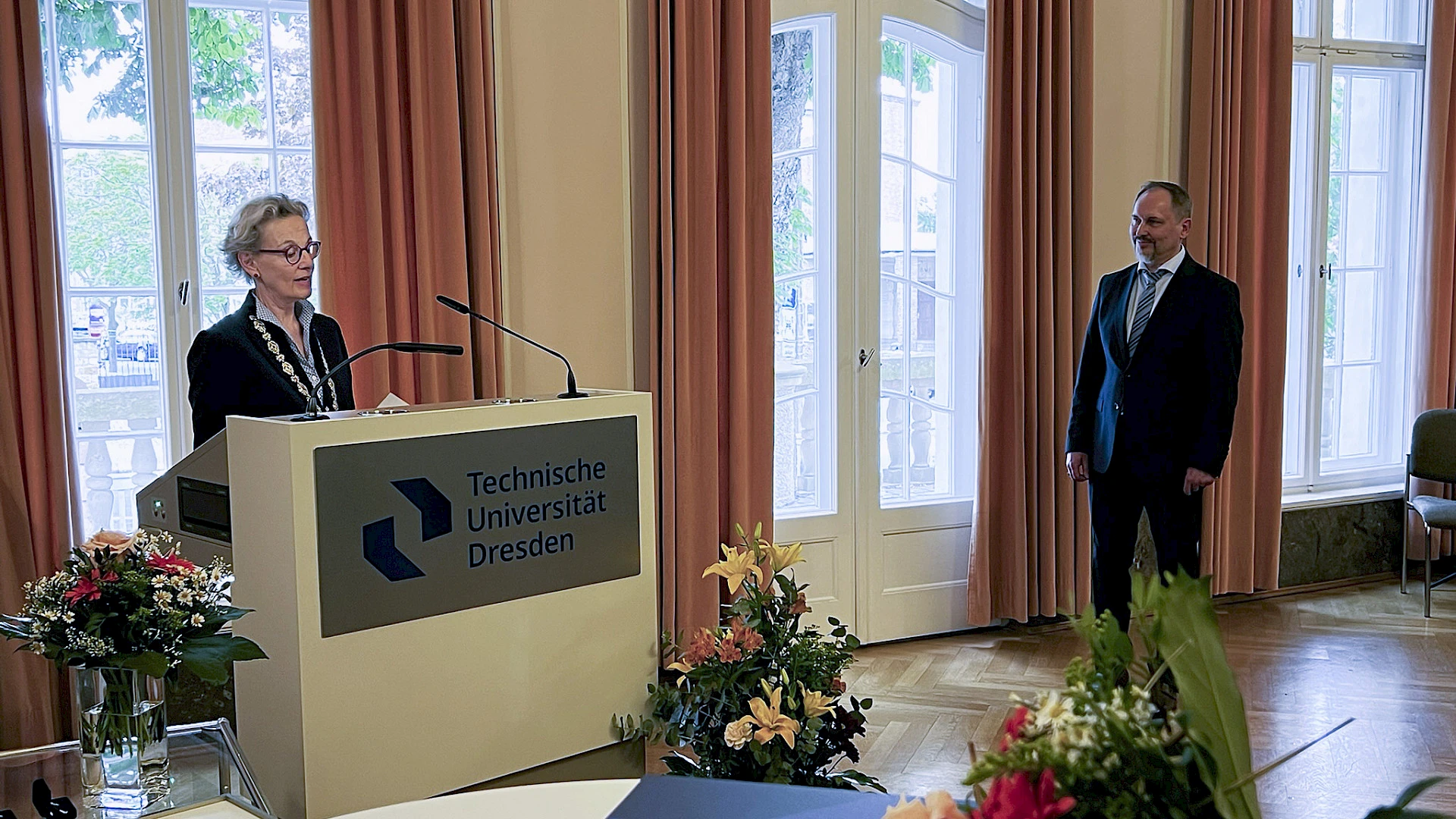 Excellent and exceptional: Prof. Dr.-Ing. Viktor Mechtcherine—spokesperson for the CARE Cluster of Excellence and member of acatech, among other roles—was awarded the Badge of Honor by the President of TU Dresden. Ceremonial Address: TUD President Staudinger speaks from a lectern in the Rectorate’s banquet hall. Several large bouquets of flowers are arranged around her. Prof. Mechtcherine, the honoree, stands next to the lectern and listens to her.