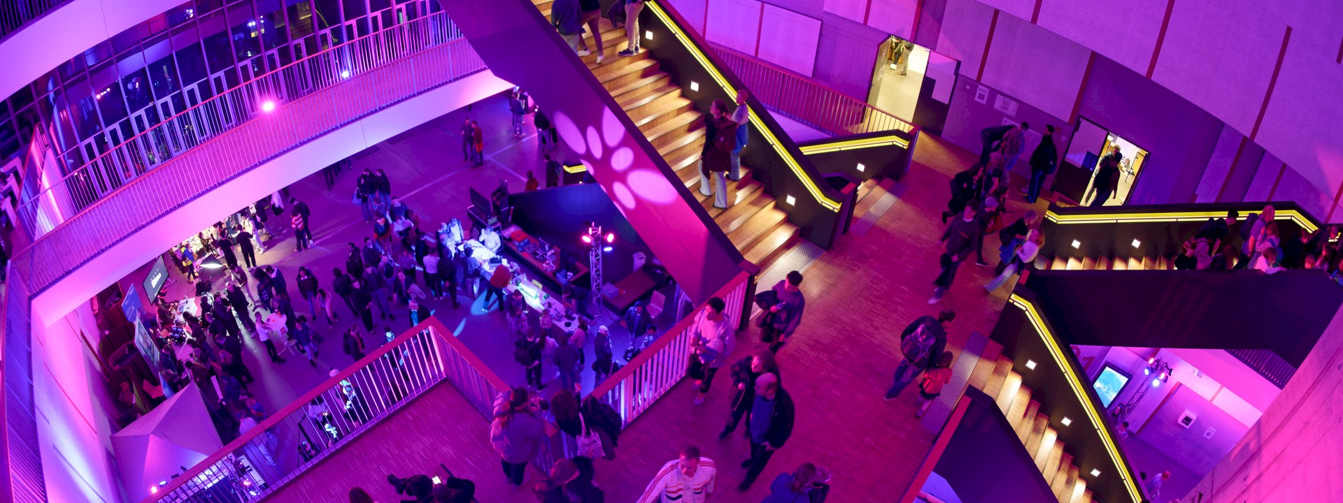 A wide-angle shot of a well-attended evening event in the modern, multi-storey foyer of a building. The entire scene is bathed in intense pink and purple light. In the centre, a large, striking wooden staircase leads upwards, with people moving along it. Numerous people are standing and walking on the main floor and the gallery levels; there are information stands and event lighting.