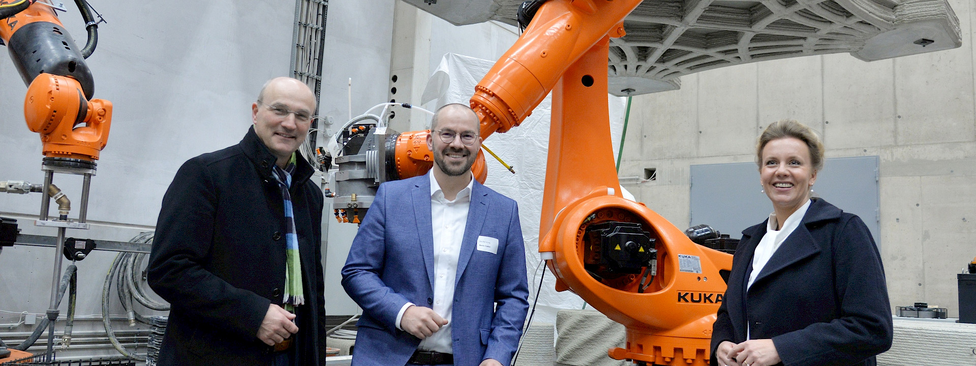 Three people – two men and a woman – standing and smiling in an industrial or laboratory setting in front of a large, orange KUKA robotic arm. In the background, a complex, 3D-printed concrete element hangs from straps.