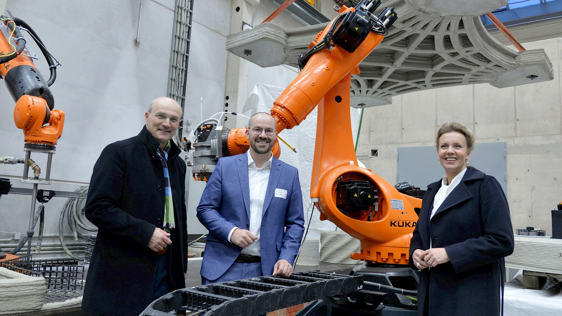 A precast concrete element suspended in the Institute of Structural Concrete hall requires only one-third of the material used in conventional fabrication. Minister Ina Brandes was welcomed to RWTH by Professor Martin Claßen (center) and the rector of RWTH Aachen University, Professor Ulrich Rüdiger. Three people – two men and a woman – standing and smiling in an industrial or laboratory setting in front of a large, orange KUKA robotic arm. In the background, a complex, 3D-printed concrete element hangs from straps.