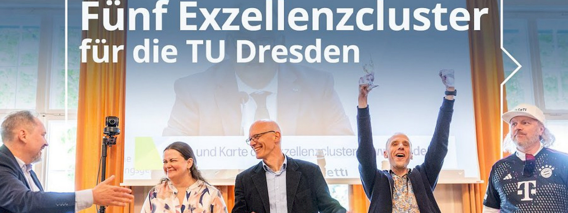 Picture of 5 cheering people in front of a video of a press conference projected on the wall