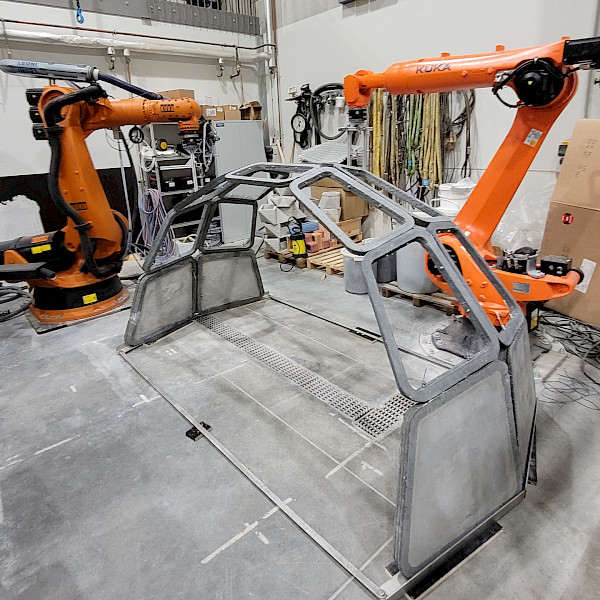 Two large, orange industrial robots in a factory hall, positioned to the left and right of an arched, grey segmented frame structure on a concrete floor.